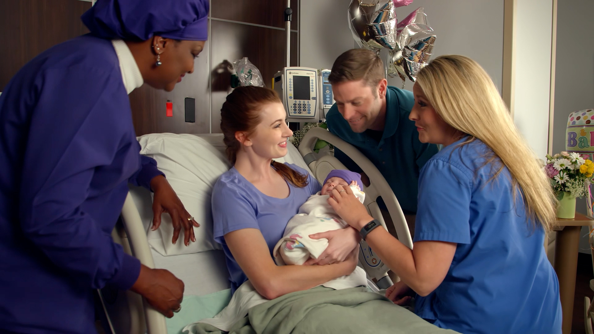 A new mother holds her newborn in the hospital bed as her partner and two nurses gather close in celebration.