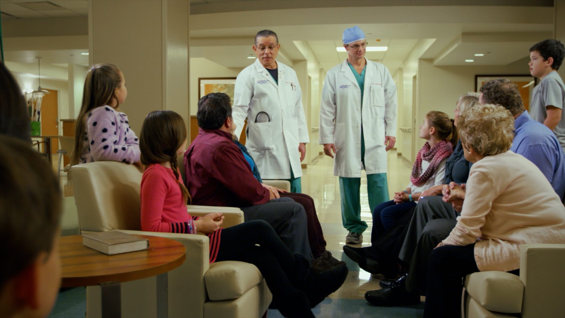 Two physicians in white coats stand together in a hospital lounge, speaking with a multi-generational family seated around them.