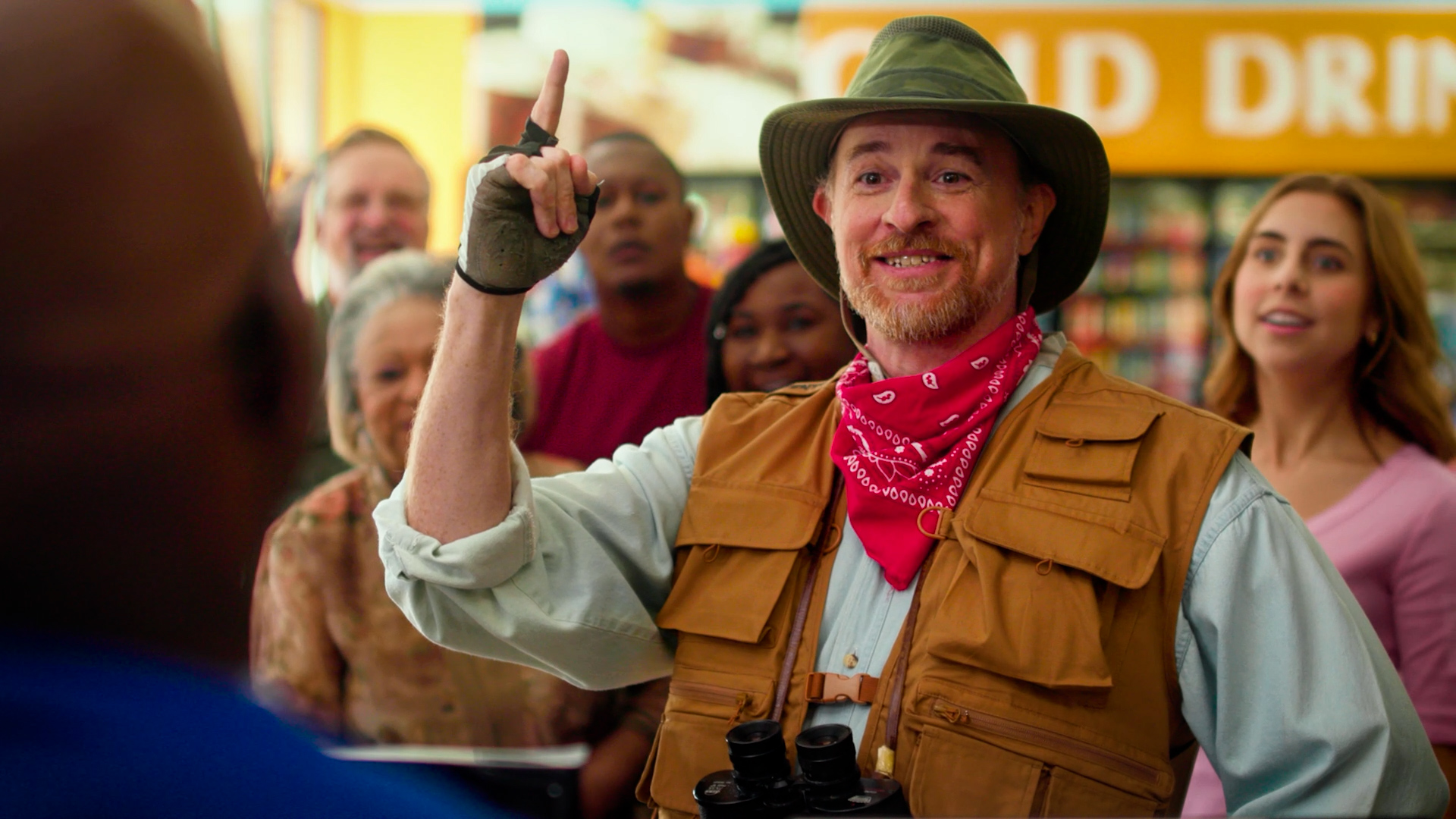 Man in a safari hat, fishing vest, red bandana, and binoculars points upward with a smile inside a convenience store aisle, surrounded by a small crowd of onlookers.