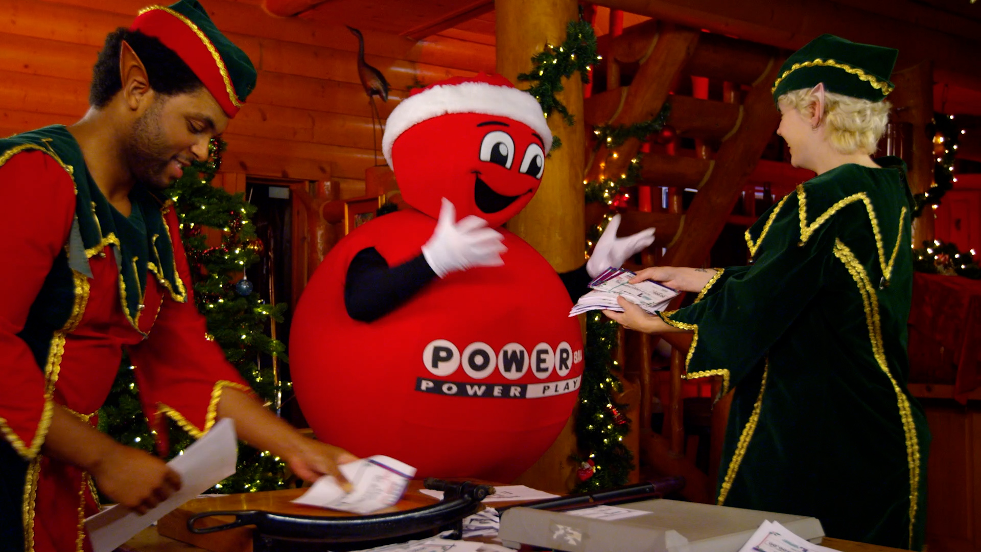 Powerball mascot in a Santa hat is handed a stack of lottery tickets by an elf, with a second elf sorting tickets on a wooden desk inside a decorated log cabin set.