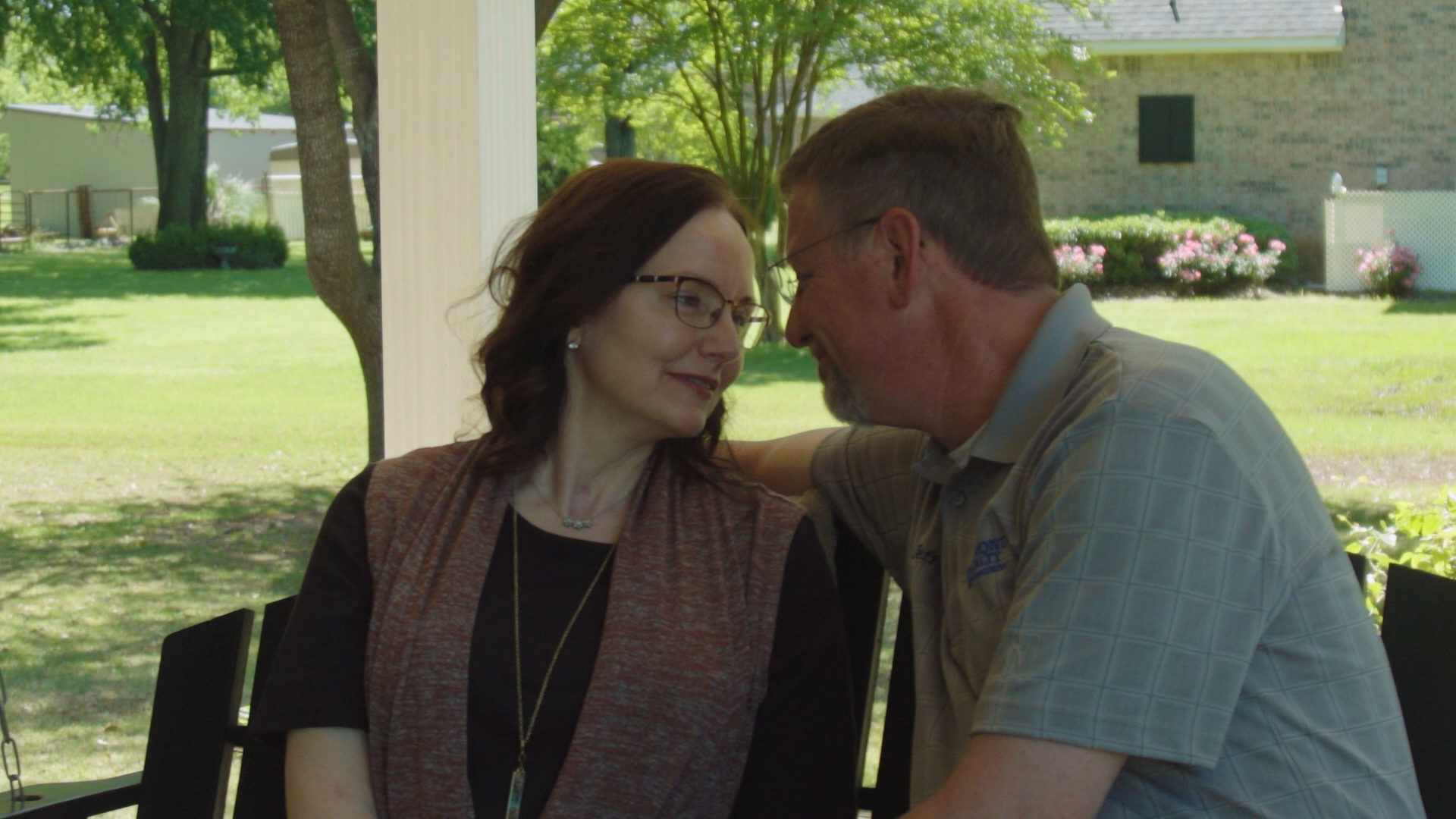 A woman and her partner sit close together outdoors on a sunlit patio, sharing a quiet, tender moment.