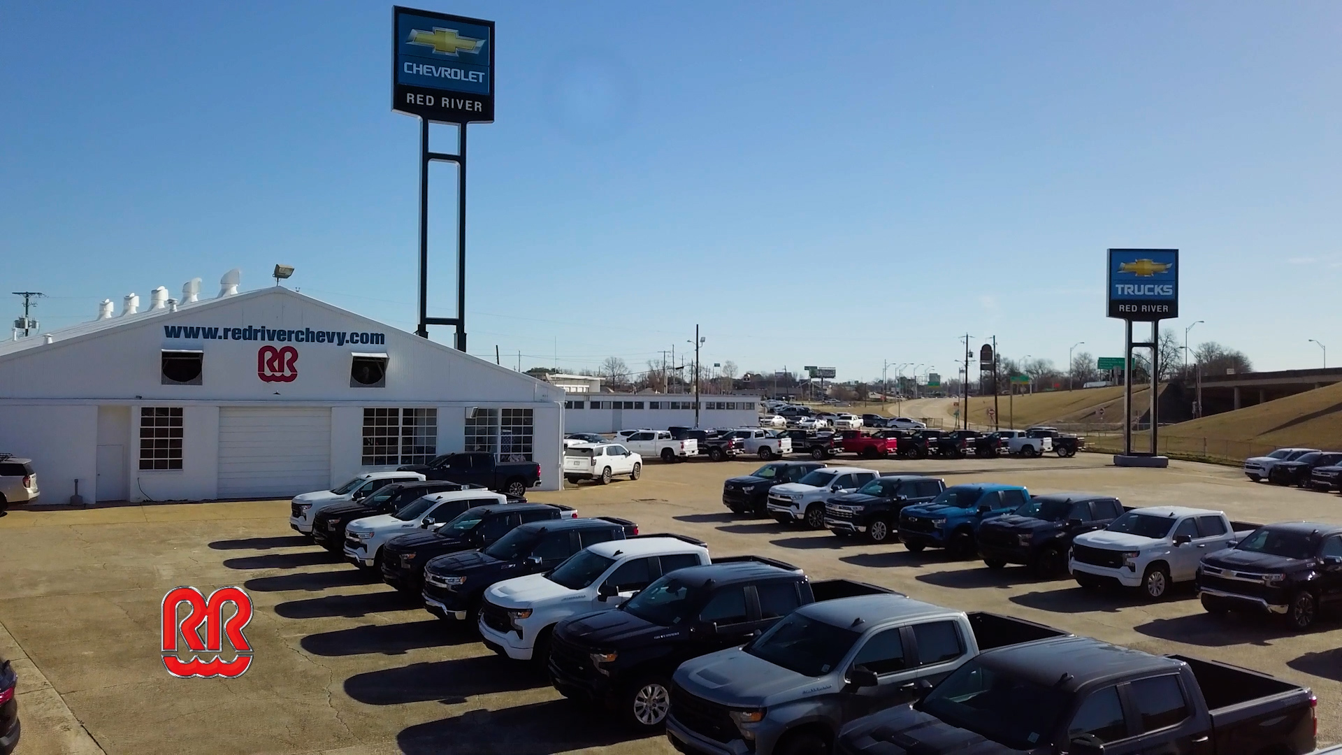 Aerial view of the Red River Chevrolet dealership lot filled with new trucks and SUVs, with branded signage and Red River logo overlay.