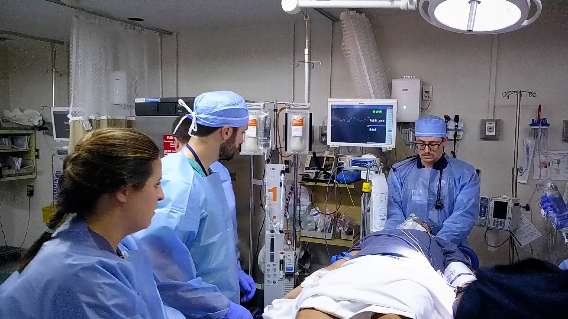 Medical team in gowns and scrubs works around a patient in Trauma Bay 1, with monitors displaying live vital signs.