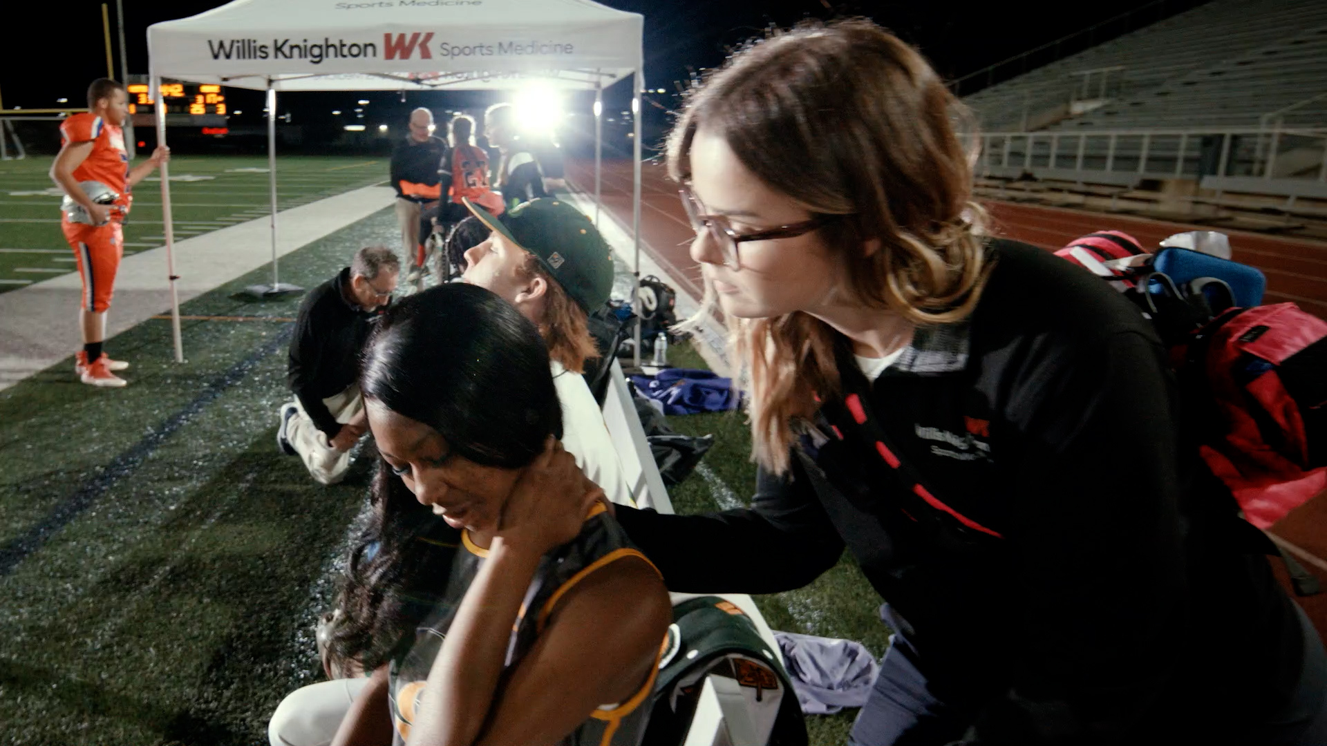 A Willis-Knighton Sports Medicine athletic trainer examines a high school athlete on the sideline of a nighttime football game, with the WK Sports Medicine tent visible in the background.